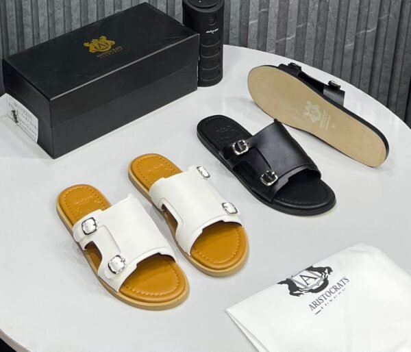 A pair of white and black double strap buckle sandals with silver buckles, accompanied by a black box and white cloth bag, showcasing the epitome of luxury footwear.