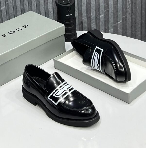 IMG-20260708-WA0044 A pair of black leather loafers with white embellishment on a white table, accompanied by a gray shoe box and a black cylindrical object.