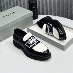 A pair of black and white patent leather loafers with contrasting strap details, displayed on a white table with a gray shoe box and a black cylindrical object in the background.
