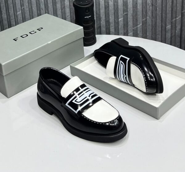 IMG-20260708-WA0048 A pair of black and white patent leather loafers with contrasting strap details, displayed on a white table with a gray shoe box and a black cylindrical object in the background.
