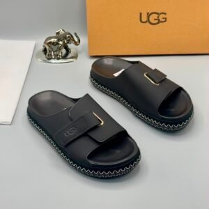 A pair of luxurious gray UGG slide sandals with gold accents and thick black and white stitched soles, displayed on a clean surface with UGG branding and packaging in the background.