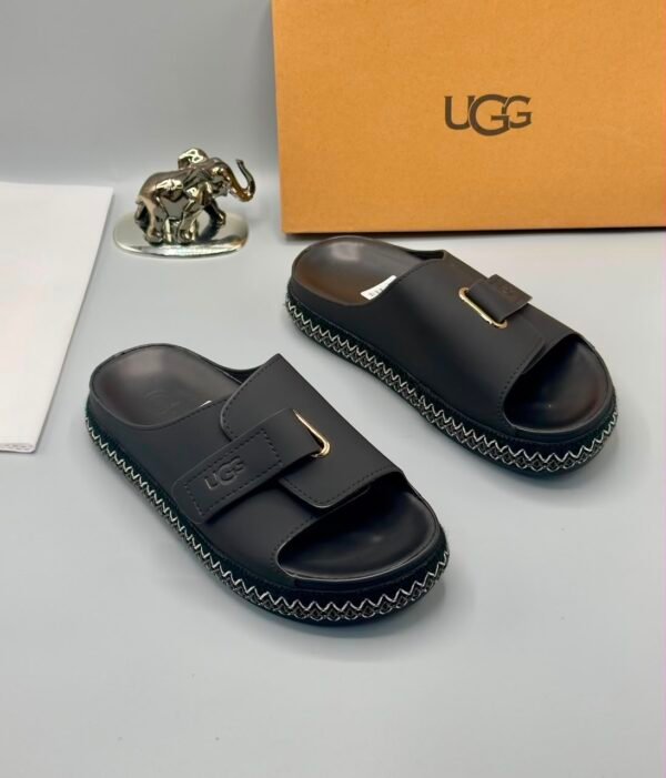 A pair of luxurious gray UGG slide sandals with gold accents and thick black and white stitched soles, displayed on a clean surface with UGG branding and packaging in the background.