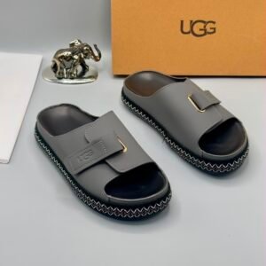 A pair of luxurious gray UGG slide sandals with gold accents and thick black and white stitched soles, displayed on a clean surface with UGG branding and packaging in the background.