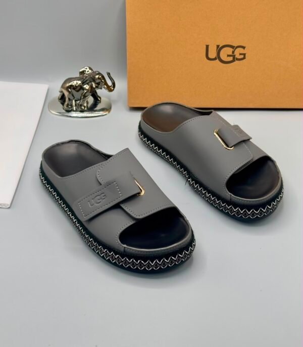 A pair of luxurious gray UGG slide sandals with gold accents and thick black and white stitched soles, displayed on a clean surface with UGG branding and packaging in the background.