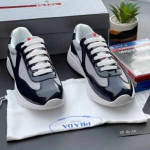 A pair of black and white Prada sneakers with white laces, placed on a white cloth bag with the Prada logo, alongside a shoe box and gaming console case.