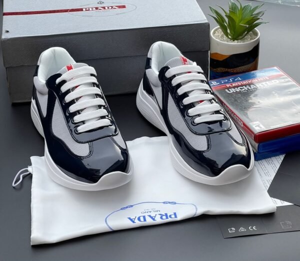 A pair of black and white Prada sneakers with white laces, placed on a white cloth bag with the Prada logo, alongside a shoe box and gaming console case.
