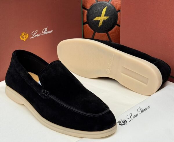 A pair of black suede loafers with cream-colored soles, displayed on a white surface with Loro Piana branding in the background.