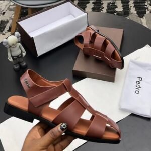 A pair of luxurious brown leather Pedro sandals with elegant strap design, presented on a stylish table with packaging and accessories.