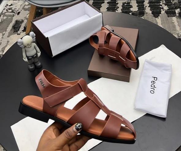 IMG-20260716-WA0007 A pair of luxurious brown leather Pedro sandals with elegant strap design, presented on a stylish table with packaging and accessories.