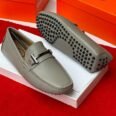 Luxury gray leather loafers with black studs on the sole, displayed on a white shoe box with an orange background.
