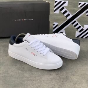 A pair of white Tommy Hilfiger sneakers with black and red logo, displayed with a black box on a concrete floor.