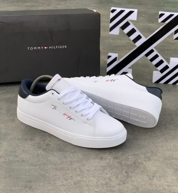 A pair of white Tommy Hilfiger sneakers with black and red logo, displayed with a black box on a concrete floor.