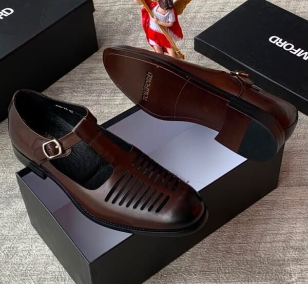 A pair of sleek black leather monk strap shoes with a silver buckle, displayed on top of an open black shoe box alongside a small angel figurine and additional closed boxes.