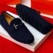 A pair of luxurious navy blue loafers with a sleek design and distinctive black buttons on the soles, presented on a white shoe box against a vibrant red background.