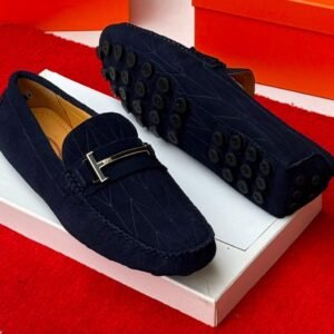 A pair of luxurious navy blue loafers with a sleek design and distinctive black buttons on the soles, presented on a white shoe box against a vibrant red background.