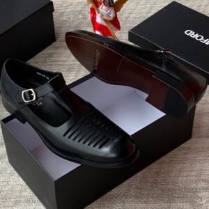 A pair of sleek black leather monk strap shoes with a silver buckle, displayed on top of an open black shoe box alongside a small angel figurine and additional closed boxes.