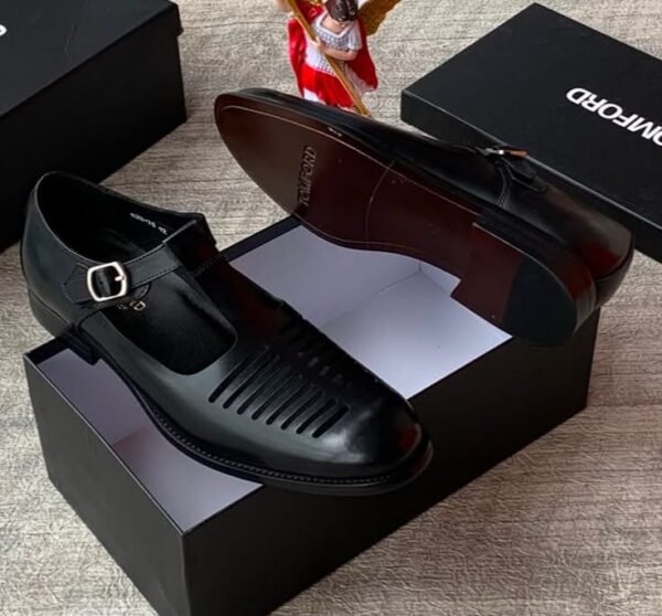 A pair of sleek black leather monk strap shoes with a silver buckle, displayed on top of an open black shoe box alongside a small angel figurine and additional closed boxes.
