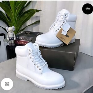 A pair of pristine white leather boots with silver laces and a chunky sole, displayed on a sleek gray surface with a shoe box, plant, and luxury brand books in the background.