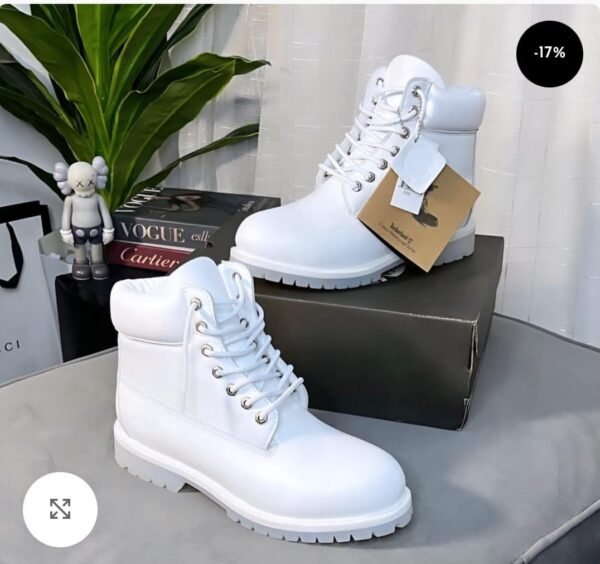 A pair of pristine white leather boots with silver laces and a chunky sole, displayed on a sleek gray surface with a shoe box, plant, and luxury brand books in the background.