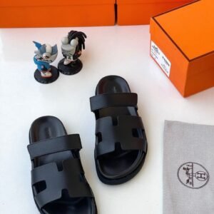 Luxury black sandals with two wide straps and a contoured footbed, displayed with orange boxes and a gray cloth bag featuring the Hermes logo.