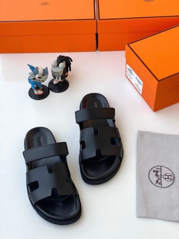 Luxury black sandals with two wide straps and a contoured footbed, displayed with orange boxes and a gray cloth bag featuring the Hermes logo.