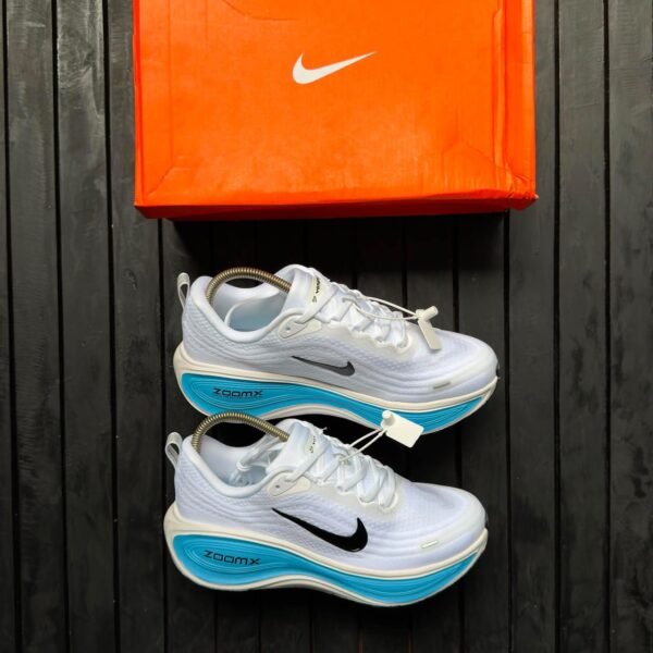 A pair of white Nike sneakers with blue soles and black Nike swoosh logo, next to an orange Nike shoe box on a dark wooden background.