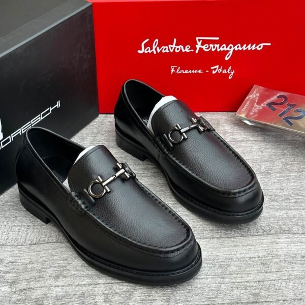 A pair of luxurious black leather loafers with metal buckles, displayed on a wooden surface with Salvatore Ferragamo box in the background.