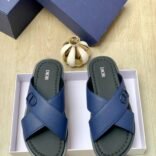A pair of blue Dior sandals with crisscross straps and gold buckle details, nestled in a white box alongside a navy blue Dior box and a gold decorative item.