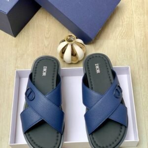 A pair of blue Dior sandals with crisscross straps and gold buckle details, nestled in a white box alongside a navy blue Dior box and a gold decorative item.