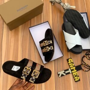 A pair of luxurious Burberry sandals, one in black with gold buckles and the other in white, displayed on a wooden surface with their respective boxes and accessories.