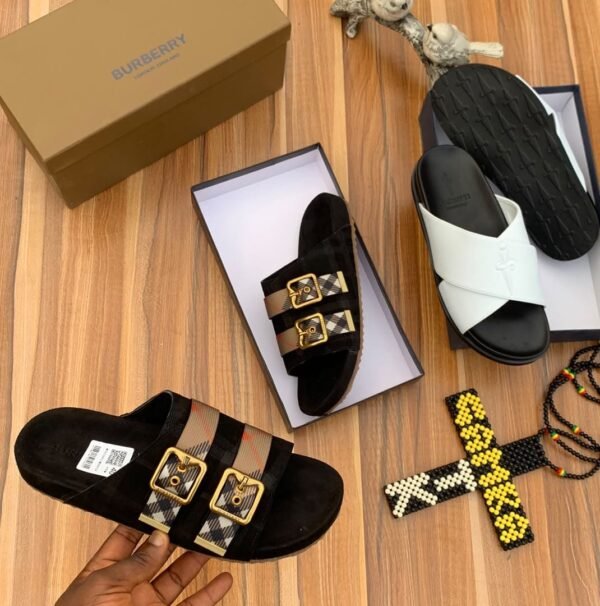 A pair of luxurious Burberry sandals, one in black with gold buckles and the other in white, displayed on a wooden surface with their respective boxes and accessories.